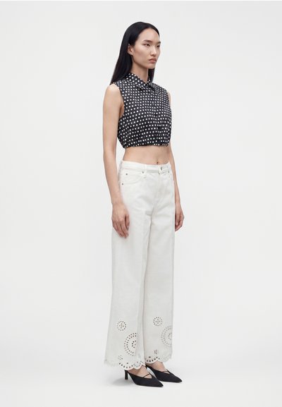 Γυναίκα με μαύρο αμάνικο crop top με πουά, λευκό παντελόνι φαρδιά μπατζάκια με σχέδιο από τρύπες και μαύρες μυτερές γόβες, στέκεται μπροστά σε λευκό φόντο.