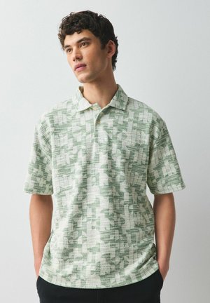 Fiatal férfi sötét, göndör hajjal, rövid ujjú zöld-fehér absztrakt mintás galléros ingben, kezét zsebre téve, kissé balra néz.