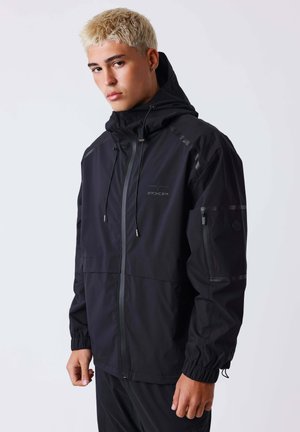 Jeune homme aux cheveux blonds courts portant une veste à capuche noire avec manches zippées et poignets élastiques sur un fond uni.