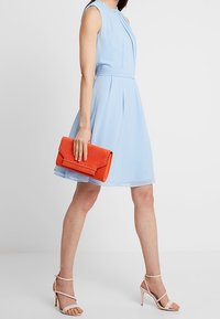 Jasnoniebieska sukienka bez rękawów z drapowanym dekoltem, w zestawieniu z jasnopomarańczową torebką kopertówką i cielistymi sandałami na obcasie.
