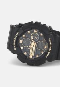 Orologio G-Shock con cinturino in resina nera, accenti dorati e quadrante testurizzato che mostra le funzioni di ora digitale e analogica. Design resistente agli urti.
