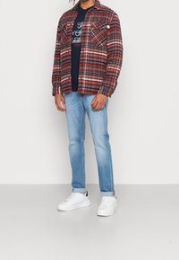 Rot-blau karierte Flanelljacke mit zwei Brusttaschen, über einem blauen T-Shirt getragen, kombiniert mit hellblauen Jeans und weißen Sneakern.