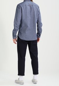Camisa de chambray azul oscuro con cuello abotonado, mangas largas y logo en el pecho, combinada con pantalones navy enrollados y zapatillas blancas.