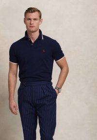 Polo azul-marinho de mangas curtas, com colarinho e detalhes em contraste. Combinado com calças azul-marinho com riscas. Relógio no pulso.