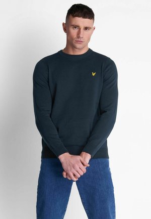 Maglione blu scuro con collo a giro e polsini e orlo ribattuti, caratterizzato da un piccolo logo di un'aquila gialla sul lato sinistro del petto. Abbinato a jeans blu.