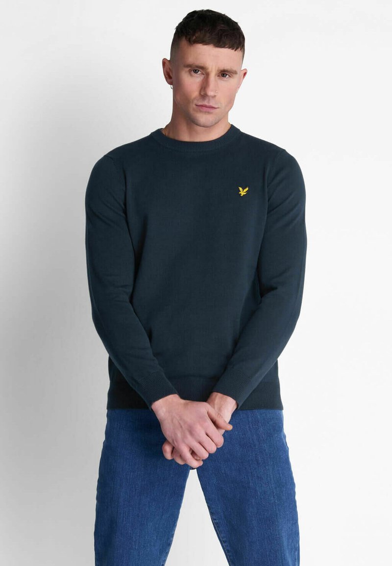 Maglione blu scuro con collo a giro e polsini e orlo ribattuti, caratterizzato da un piccolo logo di un'aquila gialla sul lato sinistro del petto. Abbinato a jeans blu.
