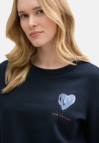 Sudadera navy con un gráfico de corazón y el texto "Tom Tailor". Hecha de una tela suave con un escote redondo y puños de canalé.