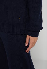 Marineblauer Strickpullover mit gerippten Bündchen und Saum. Verfügt über ein kleines goldfarbenes Logo-Detail auf der Vorderseite. Weicher Stoff und lässige Passform.