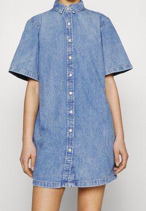 Robe chemise en denim bleu clair à manches courtes avec des boutons sur le devant, portée par une personne debout devant un fond uni.