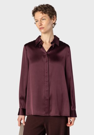 Kvinde med kort krøllet hår iført en langærmet silkeburgundy skjorte og holder en beige håndtaske mod en ensfarvet lys baggrund.