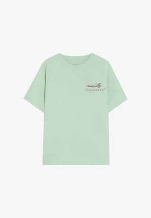 Camiseta de manga corta verde claro con un pequeño gráfico de una tabla de surf y una ola en púrpura oscuro en la parte superior izquierda del pecho.