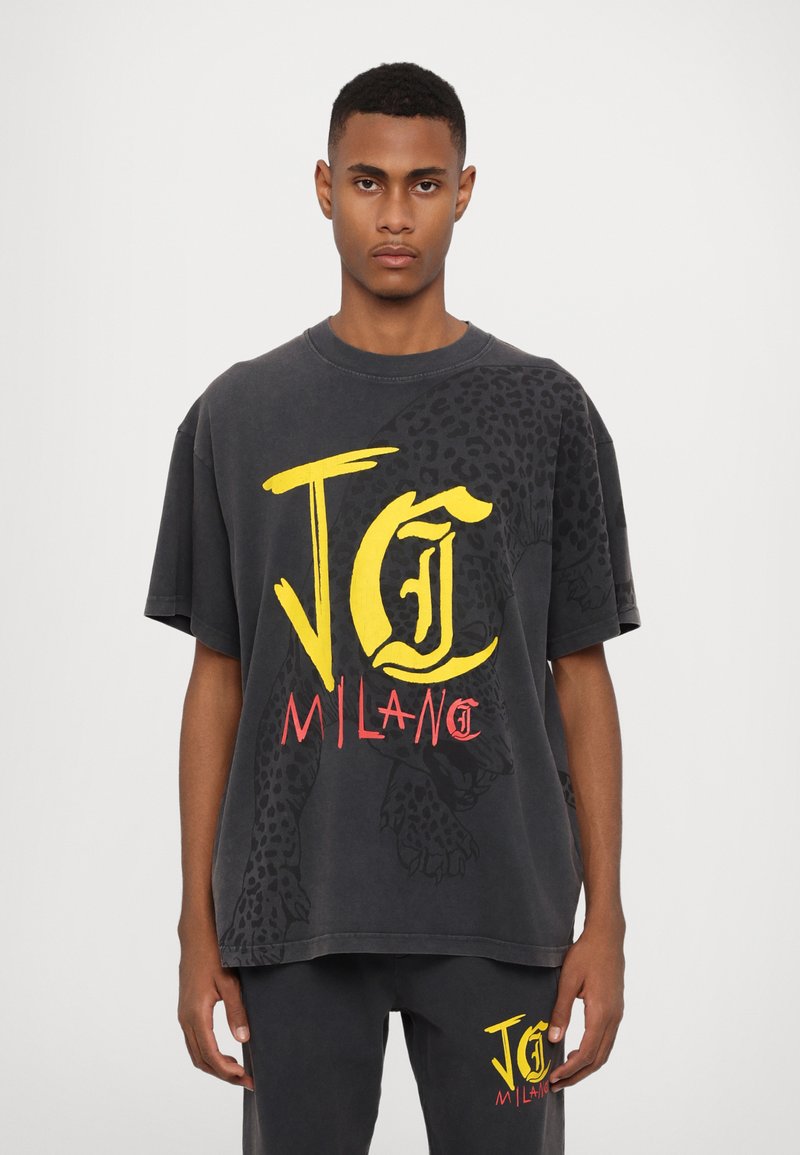 Černé oversized tričko s žlutým a červeným grafickým textem "JC MILAN" a leopardím potiskem na rameni, vyrobené z měkké bavlněné tkaniny.