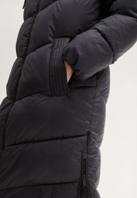 Cappotto imbottito nero realizzato in tessuto liscio e lucido, con chiusura a zip, tasche laterali e sezioni orizzontali cucite per l'isolamento.
