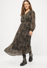Robe maxi à motifs cachemire noirs et bruns avec manches longues en voile transparent et taille smockée ; associée à des bottes noires épaisses ornées de chaînes argentées.