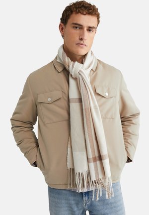 Veste beige avec deux poches poitrine, assortie à une écharpe à carreaux blanche et beige, présentant des détails en frange. Un jean bleu clair complète le look.