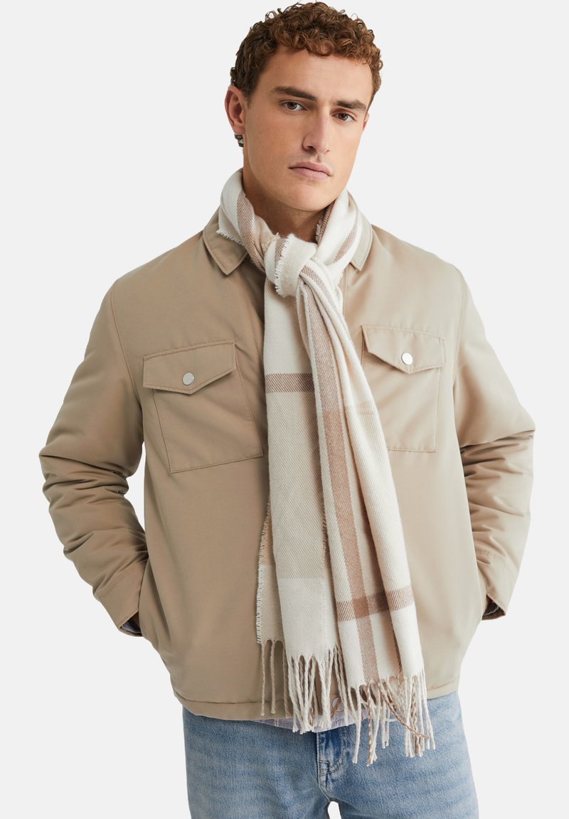 Chaqueta beige con dos bolsillos en el pecho, combinada con una bufanda de cuadros blancos y beige que presenta detalles de flecos. Unos vaqueros azul claro completan el look.