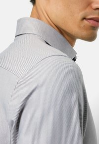 Hemd mit Kragen aus hellgrauem Stoff mit feinen, dicht beieinanderliegenden dunklen Streifen. Die Textur wirkt glatt, mit leicht tailliertem Schnitt.