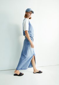 Denimrok met een zijsplit, lichtblauwe kleur, gecombineerd met een cropped wit shirt en een blauwe pet. Zwarte slide sandalen maken de outfit compleet.