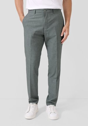 Mężczyzna w szarych eleganckich spodniach z bocznymi kieszeniami i białych trampkach, stojący na jasnoszarym, jednolitym tle.