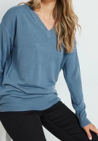 Sweter z dekoltem w serek w miękkim niebieskim materiale, z długimi rękawami i ściągaczem u dołu. Połączony z czarnymi spodniami dla eleganckiego, dopasowanego wyglądu.