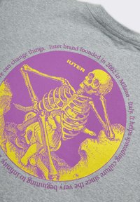 La t-shirt in cotone grigia presenta una grande grafica gialla e viola di uno scheletro. Il testo attorno al design mette in evidenza la storia del marchio Iuter.