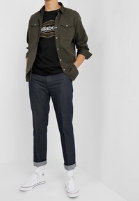 Mörk olivgrön skjorta med knappar, svart grafisk T-shirt, mörkblå avsmalnande jeans och vita canvas-sneakers med uppvikta cuffs.