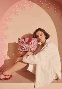 Květinová taška přes rameno v růžové barvě s červenými držadly. Hladká textura, kompaktní tvar. Modelka sedící na broskvovém pozadí.