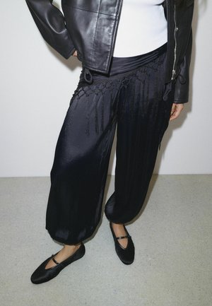 Persona che indossa pantaloni harem neri lucidi con dettagli in frange, giacca di pelle nera, top bianco e scarpe Mary Jane nere, in piedi all'interno.