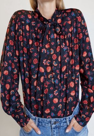 Vrouw draagt een zwarte blouse met rood en blauw bloemenpatroon en een strikje aan de kraag, ingestopt in lichtblauwe spijkerbroek.