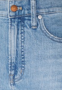 Ljusblå denimjeans har en metallknapp och nitar, med sömdetaljer längs midjebandet och fickorna. Texturen är slät och något bleknad.
