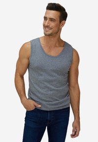 Uomo sorridente che indossa una canotta grigia senza maniche e jeans blu scuro, in piedi con una mano in tasca su sfondo bianco.
