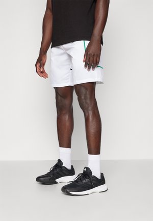 Completo sportivo maschile composto da pantaloncini bianchi con dettagli verdi, maglietta nera e scarpe da ginnastica nere con suole bianche.