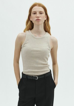Young woman with long red hair wearing a beige sleeveless top and black pants with hands in pockets against a neutral background.