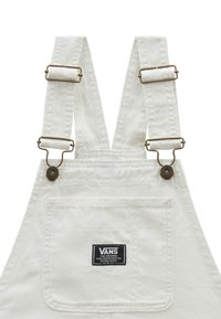Witte katoenen overalls met verstelbare messing hardware, voorzien van een voorzak en een zwart Vans logo etiket op de achterkant.