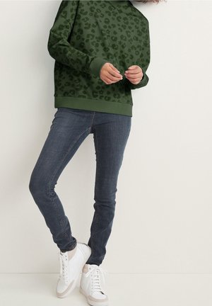 Personne portant un sweat-shirt vert à imprimé léopard, un jean skinny foncé et des baskets blanches, debout devant un fond blanc uni.