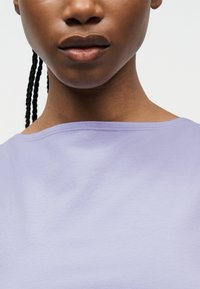 Κοντινό πλάνο ενός ατόμου με σκούρο δέρμα, πλεξούδες στα μαλλιά, που φοράει γαλάζιο μοβ τοπ με φαρδιά λαιμόκοψη, με απλό λευκό φόντο.