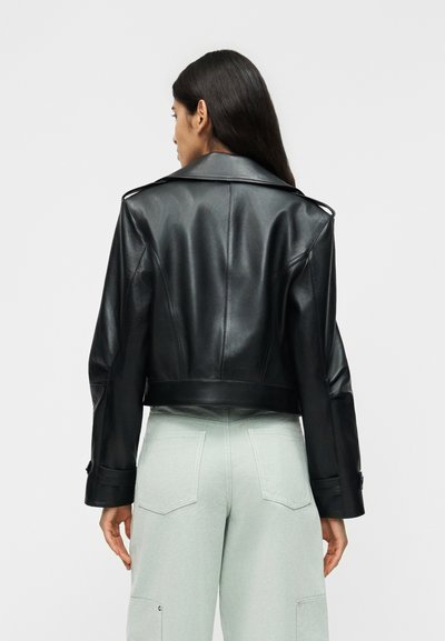 Γυναίκα με μακριά σκούρα μαλλιά που φοράει μαύρο δερμάτινο μπουφάν και ανοιχτόχρωμο παντελόνι, όρθια με την πλάτη προς το μέρος μας μπροστά σε ένα απλό φόντο.