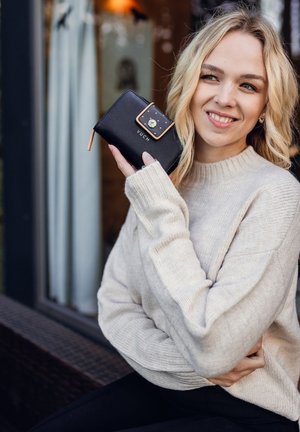 Schwarze VUCH-Brieftasche mit einem goldakzentuierten Deckel, der ein florales Design und einen Reißverschlussverschluss aufweist, gehalten von einer Person in einem cremefarbenen Pullover.
