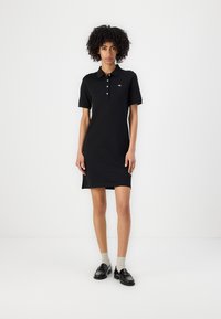 Rochie polo neagră cu mâneci scurte, material din bumbac, închidere cu trei butoane, slituri laterale și detaliu discret cu logo pe piept. Asortată cu pantofi loafer și șosete.