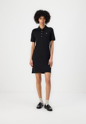 Rochie polo neagră cu mâneci scurte, material din bumbac, închidere cu trei butoane, slituri laterale și detaliu discret cu logo pe piept. Asortată cu pantofi loafer și șosete.