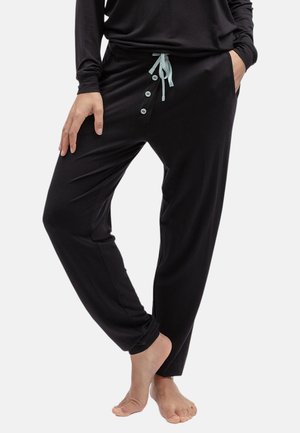 Person trägt schwarze Freizeithose mit Frontknöpfen und hellblauem Kordelzug, steht barfuß mit einer Hand in der Tasche.