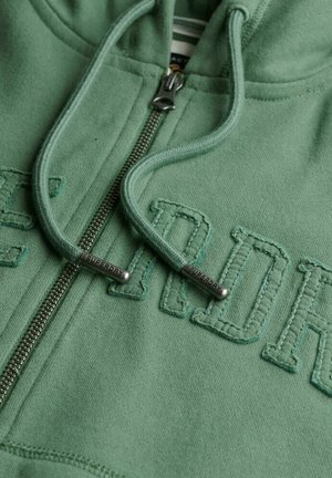 Veste à capuche zippée verte en tissu doux, avec une fermeture éclair à l'avant et une broderie texturée "SUPERDRY". Comprend un cordon de serrage avec des embouts métalliques.