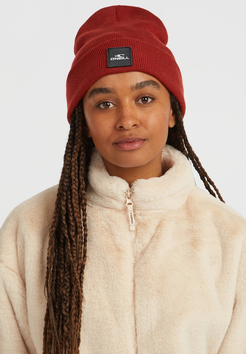 Gorro de punto acanalado rojo con un parche de logo tejido negro, combinado con una chaqueta de forro polar beige claro que cuenta con cierre de cremallera y cuello alto.