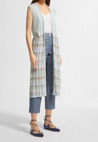 Gilet lavorato a maglia di colore azzurro chiaro con motivi a zigzag, indossato sopra una maglietta bianca e pantaloni grigi a tre quarti. La modella indossa delle scarpe blu con cinturini neri.