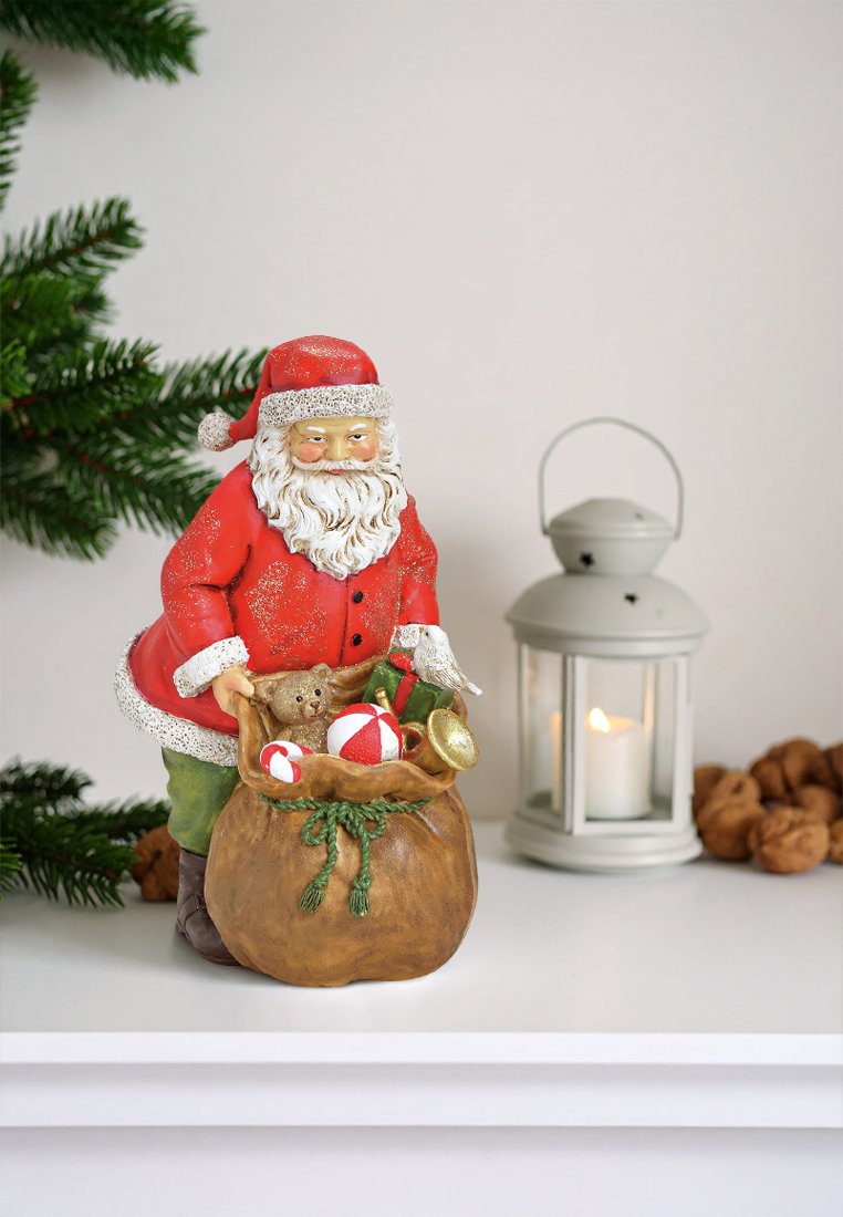 Figuur van de Kerstman in rode kleding met witte randen, die een bruine zak met snoep en een teddybeer vasthoudt. Lantaarn op de achtergrond.