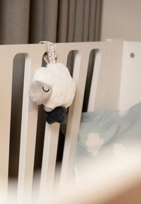 Pluche schaapje met een wit, pluizig lijf, een grijs gezicht en donkere poten, bevestigd aan een ledikantje met een lint met stippen. Zacht, textuurrijk materiaal.