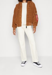 Hnědá fleecová bomber bunda s kapucí, zipovým zapínáním a bočními kapsami. V kombinaci s bílými rovnými kalhotami a černými teniskami.