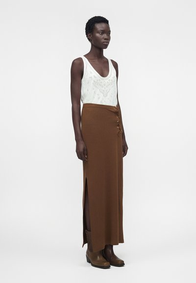 Γυναίκα με κοντά μαλλιά που φορά λευκό αμάνικο κορμάκι, καφέ μακριά φούστα με ψηλή μέση και πλευρικό σκίσιμο, και καφέ μποτάκια αστραγάλου, στέκεται πλαγίως.