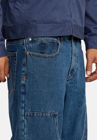 Jeans en denim dans un bleu moyen avec des coutures visibles, une broderie de rose sur la poche arrière et un design ample.