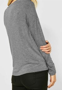 Graues Langarmoberteil mit lockerem Schnitt, runder Ausschnitt und leicht abgesenkten Schultern. Weicher, strukturiert Stoff; seitliche Schlitze am Saum.